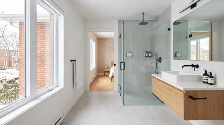 Salle de bain rénovée moderne et lumineuse intégrée harmonieusement dans une maison montréalaise, avec douche vitrée, meuble-lavabo suspendu contemporain et carrelage neutre, baignée de lumière naturelle